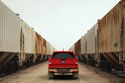 A red Ford Ranger pickup truck viewed from the rear with an expanded Sawtooth STRETCH truck bed cover, positioned in the center of a dirt track between long rows of cargo train cars under a pale sky.