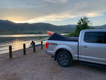 A silver Ford F-150 pickup truck parked on a gravel shore beside a lake at sunset, with a black tonneau cover and an orange kayak visible in the bed. Mountains and trees are in the background, and a person sits near the water.