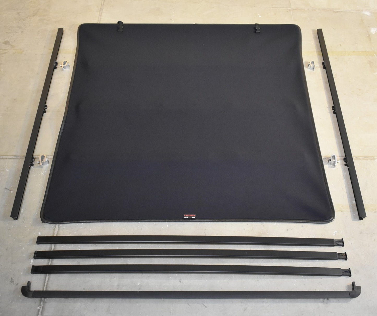 Black Sawtooth STRETCH truck bed cover soft top cover with four mounting rails and four support bars, laid out on a light concrete floor.