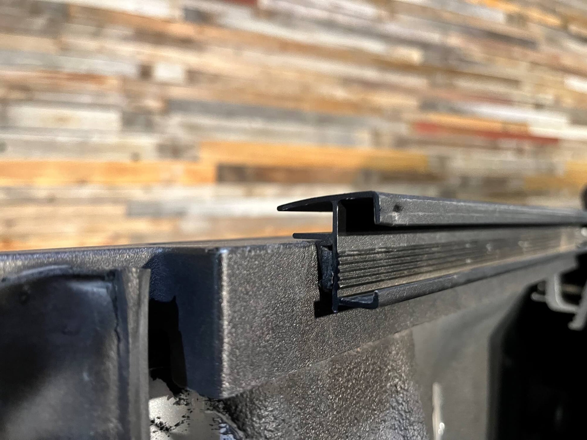 Close-up of a Sawtooth STRETCH truck bed cover powder coated black aluminum side rail with a raised lip, shown against a blurred wood plank background.