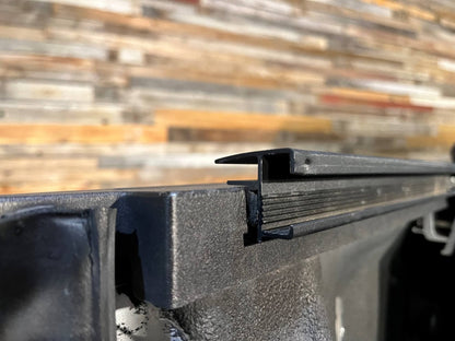 Close-up of a Sawtooth STRETCH truck bed cover powder coated black aluminum side rail with a raised lip, shown against a blurred wood plank background.