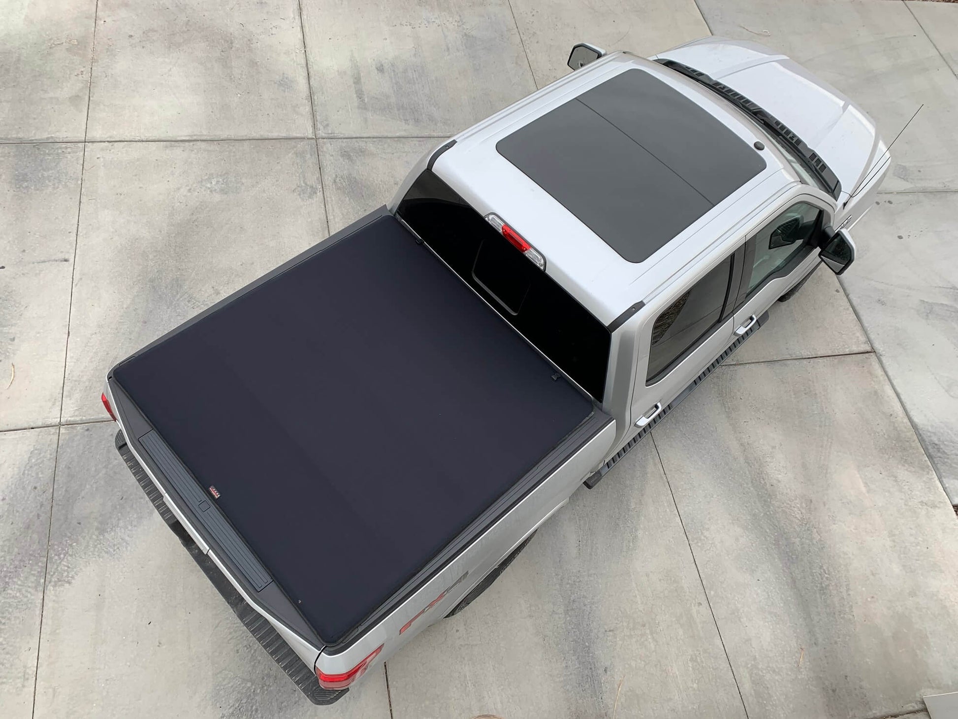 A silver pickup truck shown from a high-angle overhead view, fitted with a dark Sawtooth Expandable tonneau cover on the bed, parked on a light gray concrete surface.