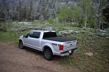 A silver Ford F-150 pickup truck with a black Sawtooth STRETCH A.C.M.S. tonneau cover is parked on a gravel path in a wooded outdoor setting with trees and shrubs in the background.
