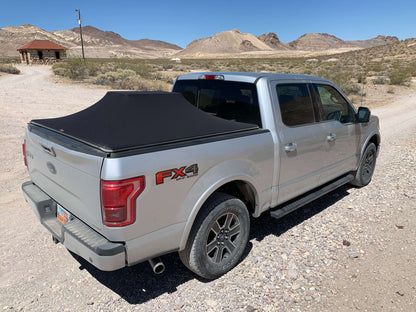 A silver Ford F-150 pickup truck parked outdoors on a gravel desert road, shown from the rear three-quarter angle with a black Sawtooth STRETCH A.C.M.S. tonneau cover over the bed. The background features desert mountains, shrubs, and a clear blue sky.