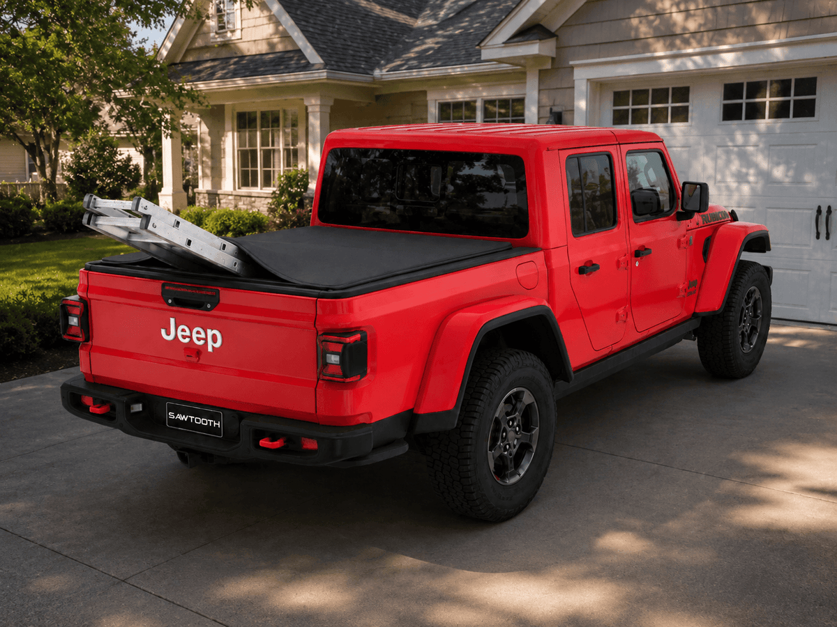A red Jeep Gladiator pickup truck parked in a residential driveway, shown from a rear three-quarter angle. The truck is equipped with a black tonneau cover and a silver ladder rack extending from the bed.