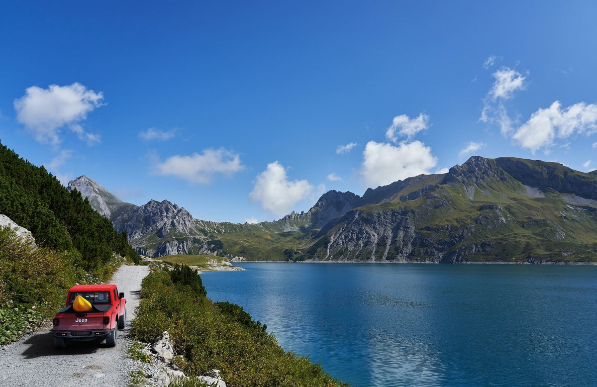 A scenic mountain lake landscape with a red pickup truck parked on a road beside the water under a bright blue sky with scattered clouds.