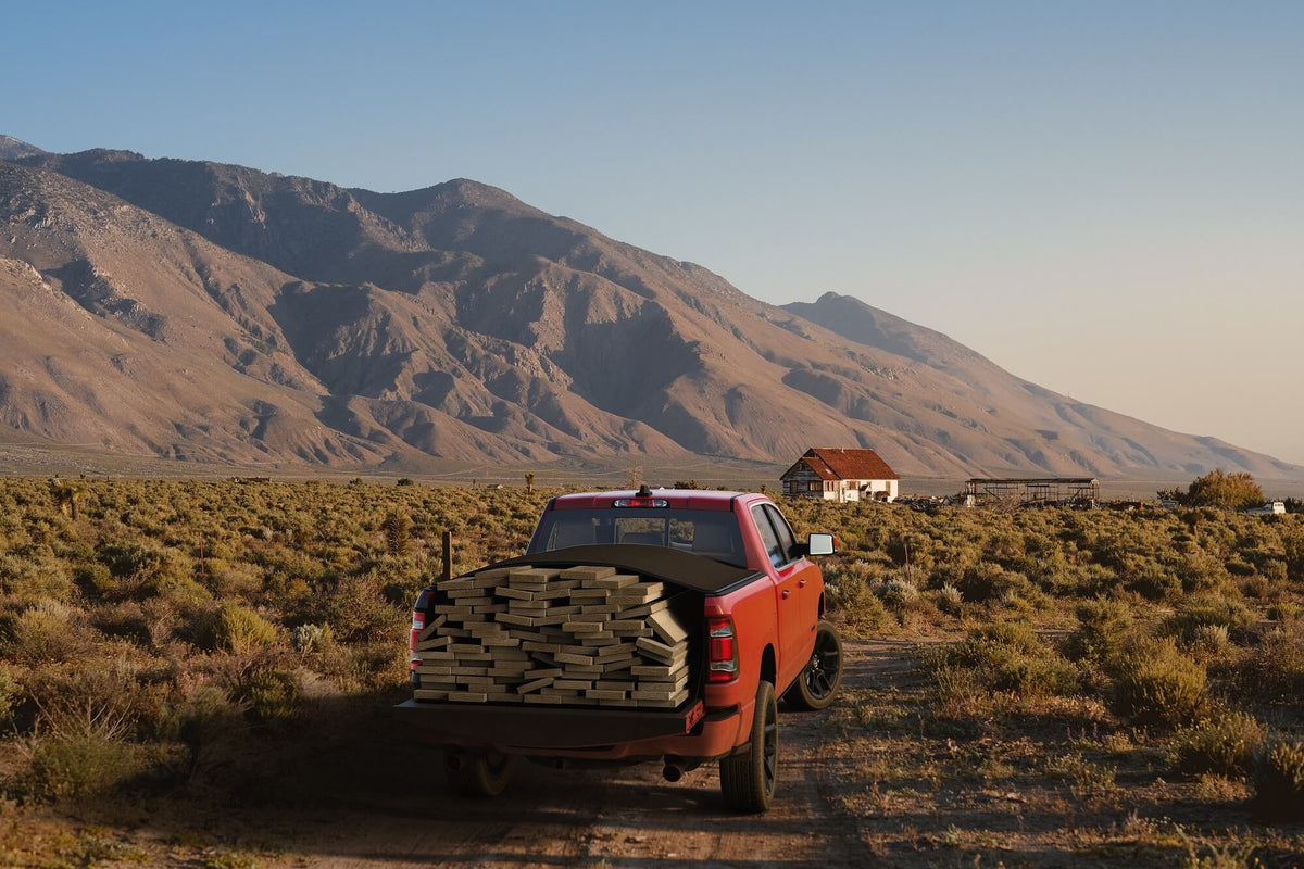 An orange pickup truck loaded with stacked lumber drives down a dirt road through a dry, scrub-covered landscape with large mountains in the background.