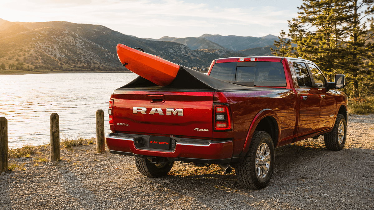 A red Ram pickup truck parked outdoors beside a lake with mountains in the background, carrying an orange kayak in the bed.