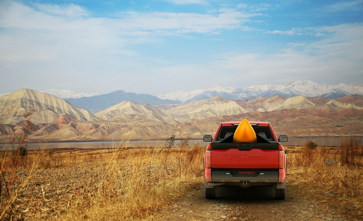 A red pickup truck viewed from behind, parked on a dirt road in a dry grassy field with layered mountains and a blue sky in the background. A bright yellow kayak rests in the bed of the truck.