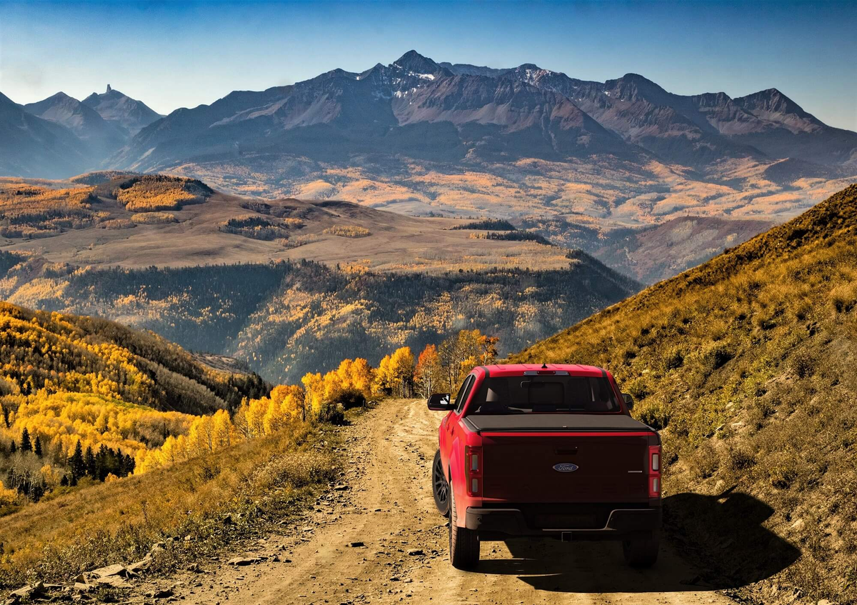 A red Ford pickup truck is shown from the rear, driving on a dirt road through a scenic mountain landscape with autumn yellow trees and blue sky.