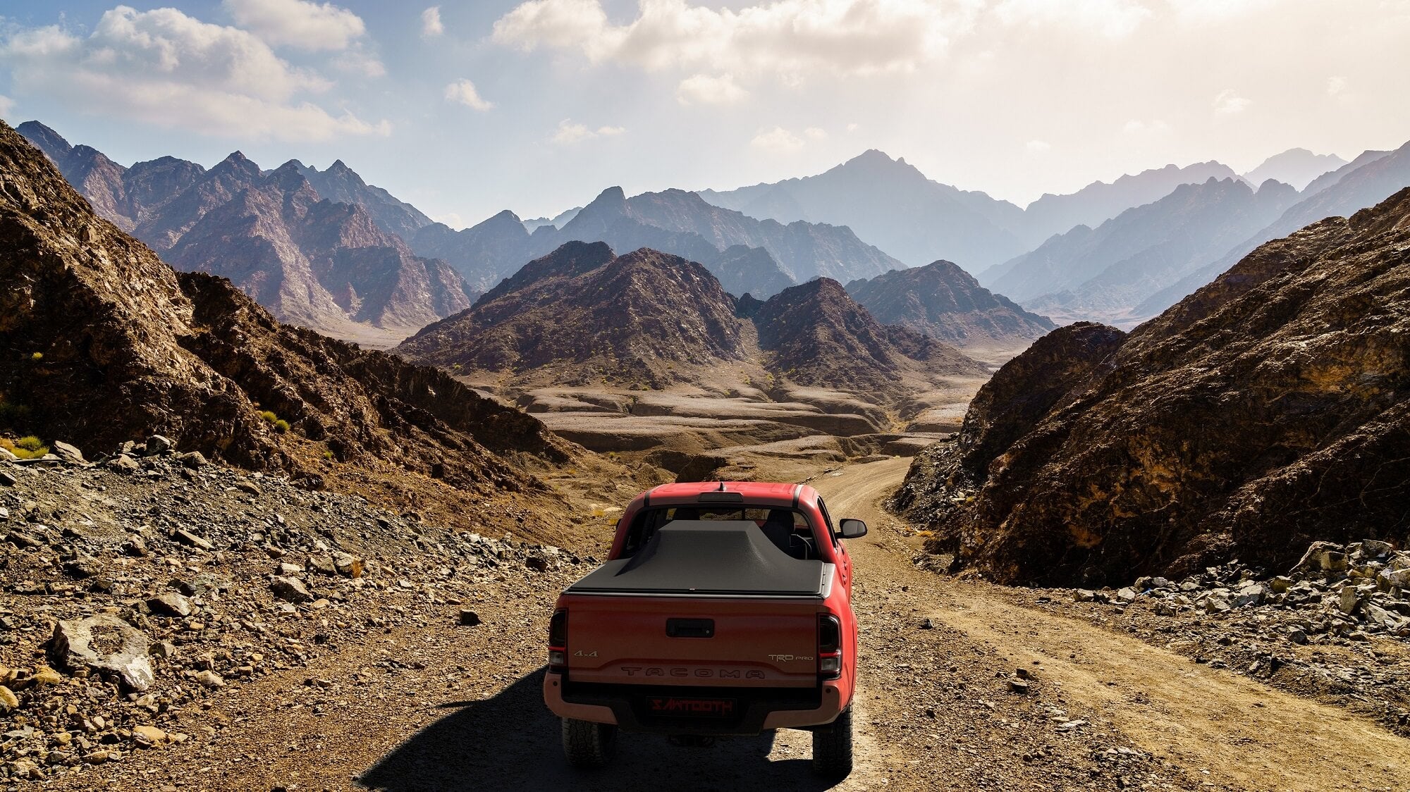 A red pickup truck viewed from behind driving on a dusty mountain road between rugged rocky peaks under a cloudy sky.