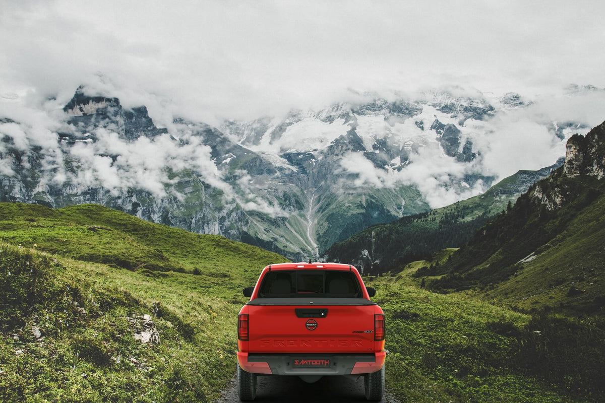A red pickup truck viewed from the rear, driving on a narrow road through a lush green mountain valley with snow-capped peaks and clouds overhead.